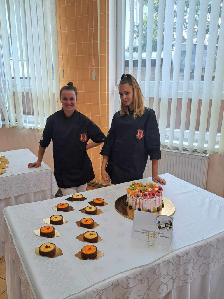 Két fekete szakácskabátos ember áll egy asztal mögött, ahol különféle desszerteket kínálnak. A bal oldalon több kis torta, a jobb oldalon pedig egy kerek tálcán díszített nagy torta. Az asztal fehér ruhával van lefedve egy olyan szobában, ahol ablakok és függőleges redőnyök állnak a háttérben.