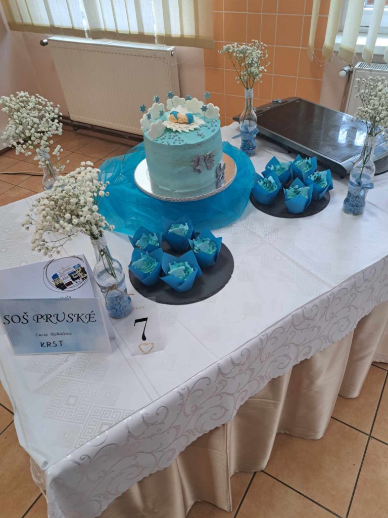 Az ünnepi terített asztalon egy kék tüll alapra helyezett menta zöld torta csillagdíszekkel és fehér virágokkal a tetején. A tortát kék virág formájú cupcakes veszi körül. Az asztalt fehér virágvázák és "SOŠ PRUŠKÉ" feliratú kis kártya díszíti.