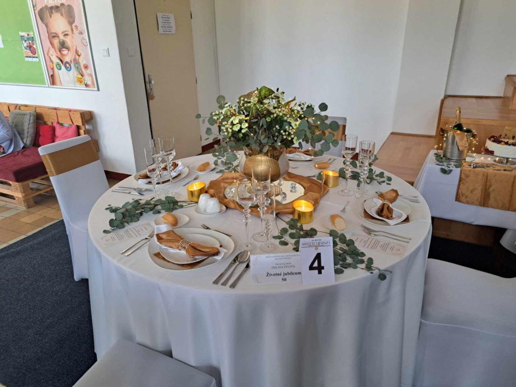 A kerek esküvői fogadóasztal elegánsan megterített fehér ágyneművel, arany díszítéssel és zöld lombozattal. A középpontban egy virágkompozíció található meggyújtott gyertyákkal. Az asztal 4-es számmal van ellátva, és a szépen elrendezett terítékek közé tartozik az étlap, a szalvéta és a poharak.
