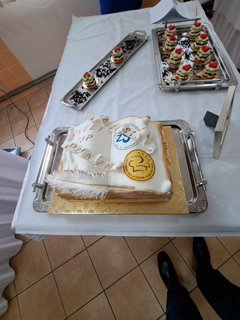 A téglalap alakú torta fehér cukormázzal és logóval a tetején látható az asztalon. A torta írással és arany kör alakú mintával. A tortát körülvevő ezüst tálcák apró, díszített desszertekkel. Egy személy lába részben látható a kép alján.