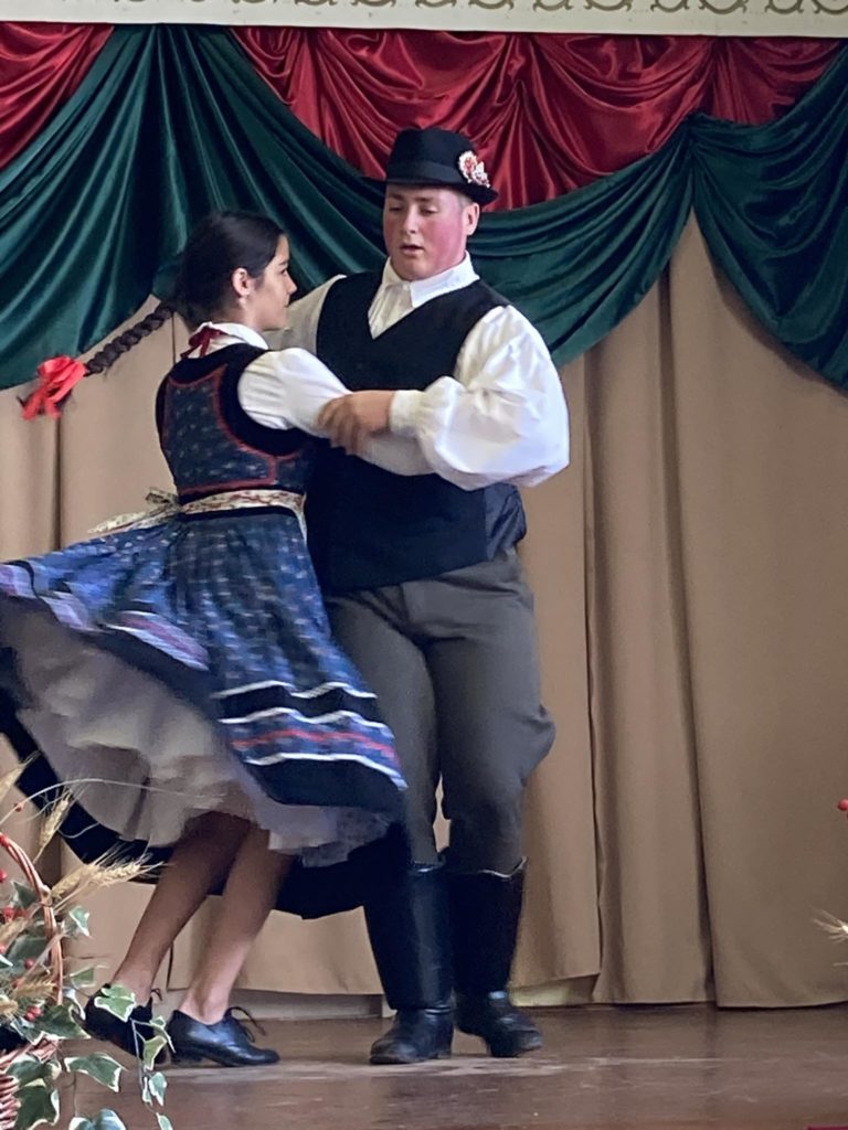 Két ember táncol hagyományos népviseletben. A nő fehér részletekkel díszített kék ruhát visel, fonott hajában piros szalaggal. A férfi fehér inget, sötét mellényt, sapkát és nadrágot visel. Vörös-zöld drapériás háttér előtt lépnek fel.