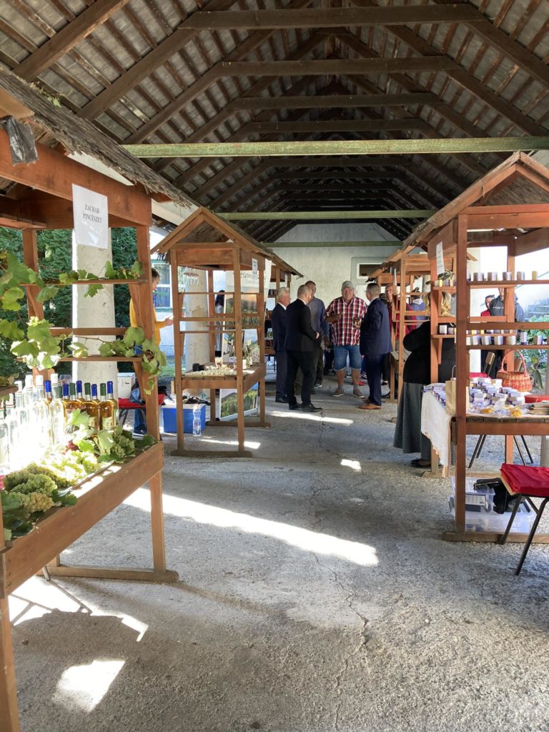 Fedett szabadtéri piac különféle árucikkeket bemutató fa standokkal. Több ember is látható, amint sétál és böngészi a tárgyakat. A piac rusztikus tetővel rendelkezik, az oldalakon elhelyezett bódék pedig palackokat, termékeket és kézműves termékeket mutatnak be.