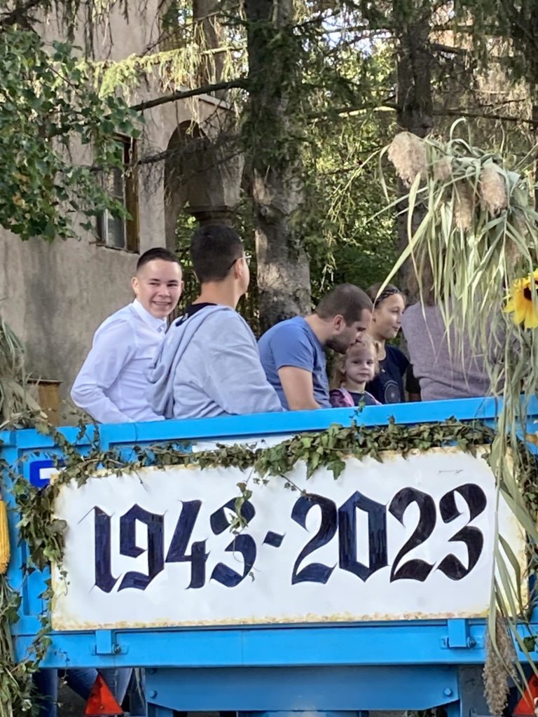 Emberek egy csoportja, köztük férfiak, nők és egy gyerek, egy díszített kék pótkocsin ül, amelyen az „1943-2023” évszám látható. A trailert növények és napraforgók díszítik, a háttérben fák láthatók.