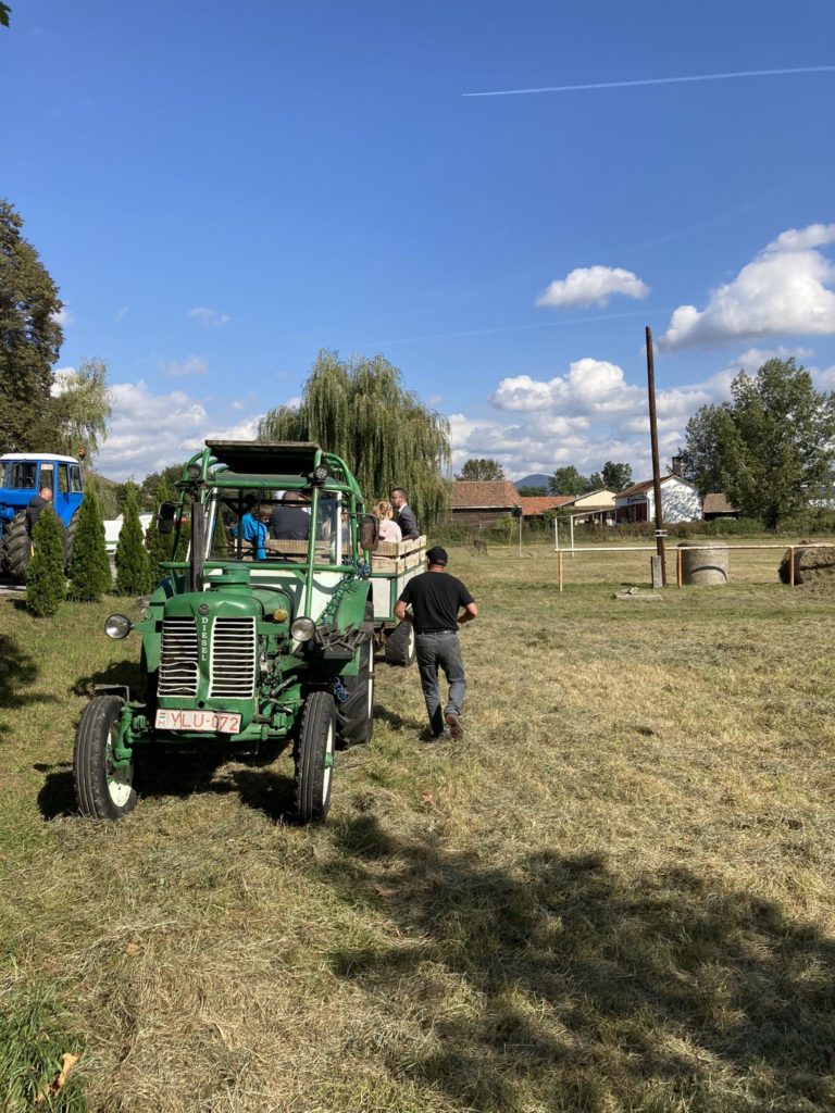 Egy személy egy zöld traktor felé sétál egy füves mezőn. Egy másik személy és egy gyerek ül a traktor hátuljában. A háttérben egy fa lógó ágakkal, néhány épület és egy kék teherautó. Az ég részben felhős, derült.