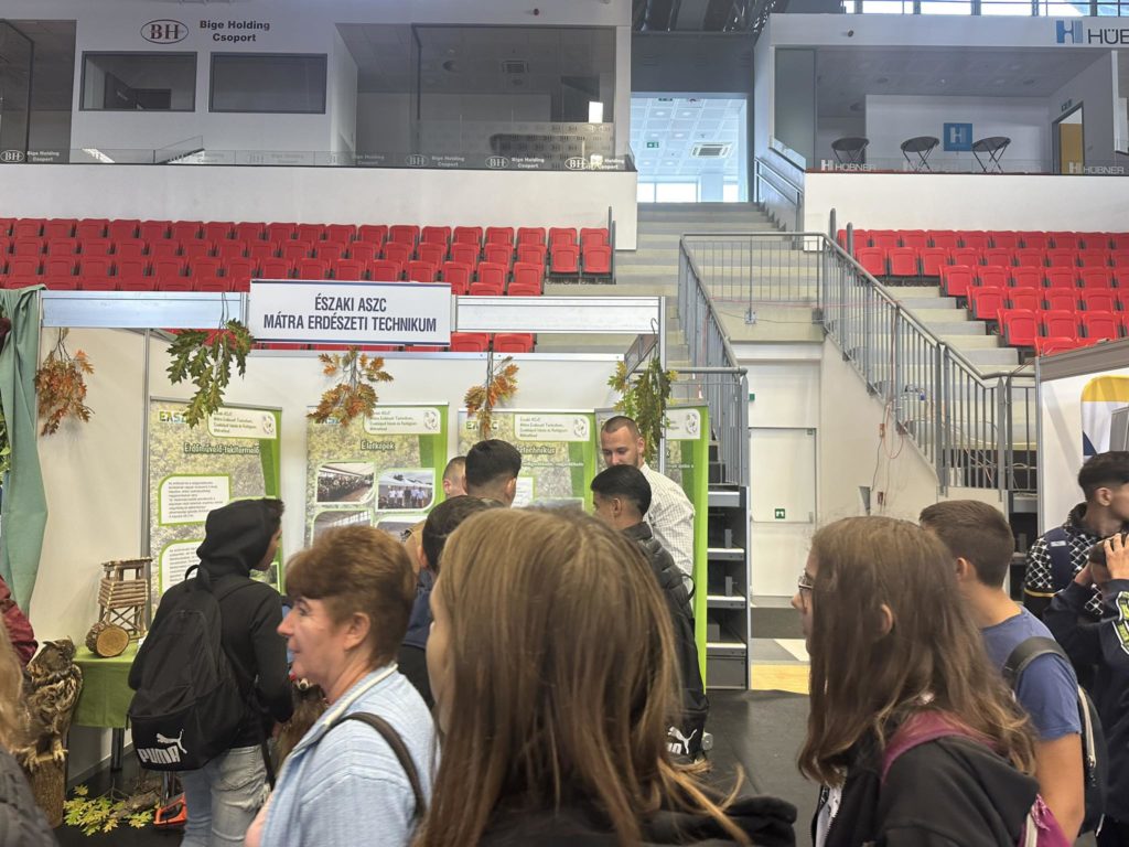 Az emberek a benti kiállítási standnál gyűltek össze. A standot természeti témájú transzparensek és különféle zöld növények díszítik. A hátteren ez olvasható: „ÉSZAKI ASZC MÁTRA ERDÉSZETI TECHNIKUM”. A térben piros ülőhelyek és lépcsők vezetnek a felső szintre.