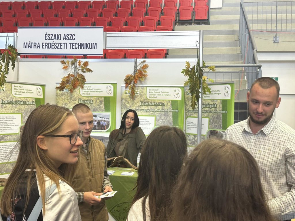 Az „Északi ASZC Mátra Erdészeti Technikum” információs standjánál egy csoport, köztük négy fiatal és egy felnőtt beszélget. A standon oktatási plakátok és őszi témájú dekorációk láthatók.