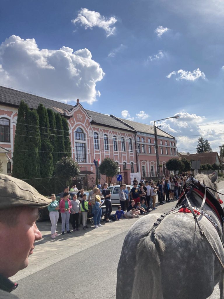 Emberek tömege áll az utcán egy nagy, díszes rózsaszín épület előtt, magas íves ablakokkal. Kék az ég, szétszórt felhők. Az előtérben egy ló feje és egy sapkát viselő személy látható részben.