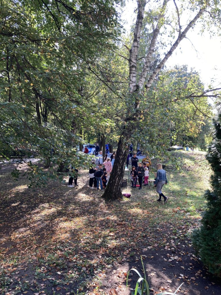 Emberek csoportja, köztük gyerekek és felnőttek, egy fa alatt gyűlik össze egy zöld fűvel és lehullott levelekkel teli parkban. A napfény átszűrődik a fákon, a háttérben pedig egy kék jármű látható. A jelenet élénknek és közösséginek tűnik.