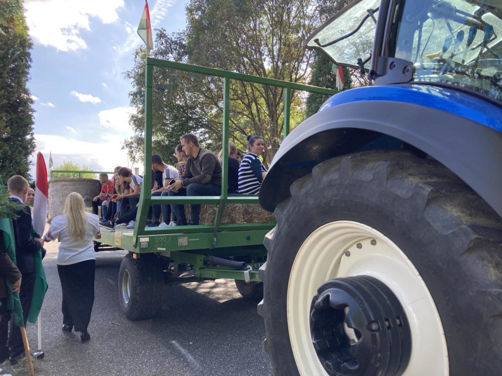 Egy nagy kék traktor egy zöld pótkocsit húz, tele szénával és emberekkel. A jelenet a szabadban, egy napsütéses napon, a háttérben fákkal. Vannak, akik zászlót tartanak, és egy fehér inges ember áll az utánfutó közelében.
