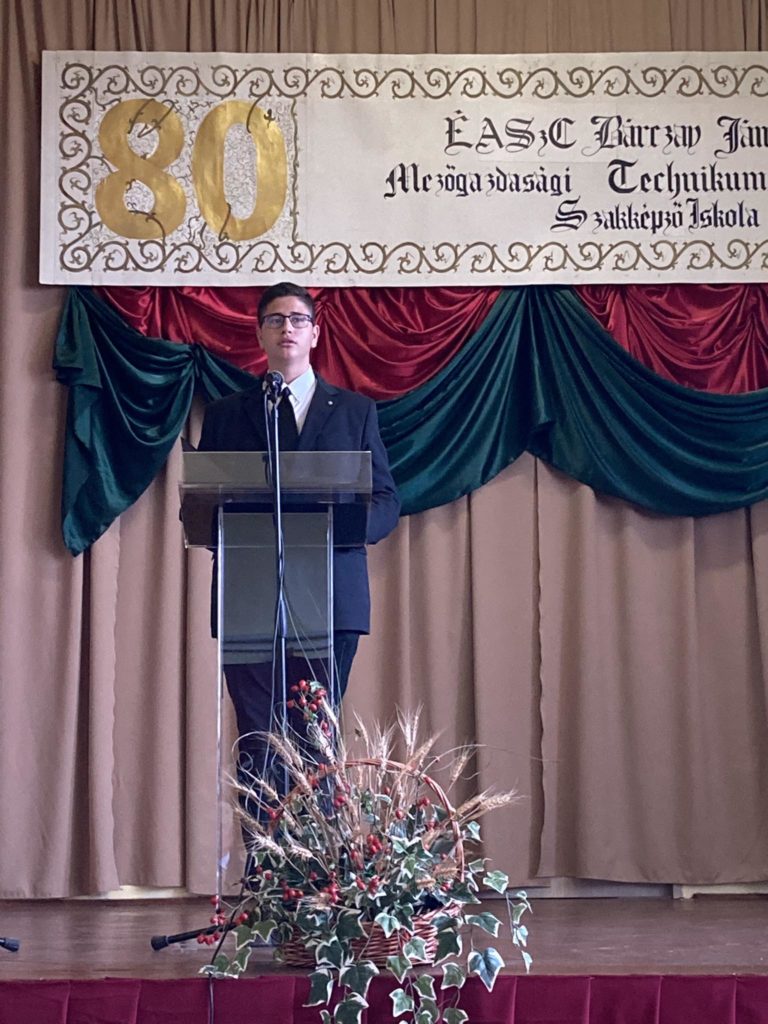Egy sötét öltönyös férfi áll az emelvényen mikrofonnal, és beszédet mond. Mögötte egy transzparens "80" arany színnel és magyar szöveggel. A színpad hátterét zöld és piros drapériák jellemzik, a pódium elején pedig virágkompozíció található.