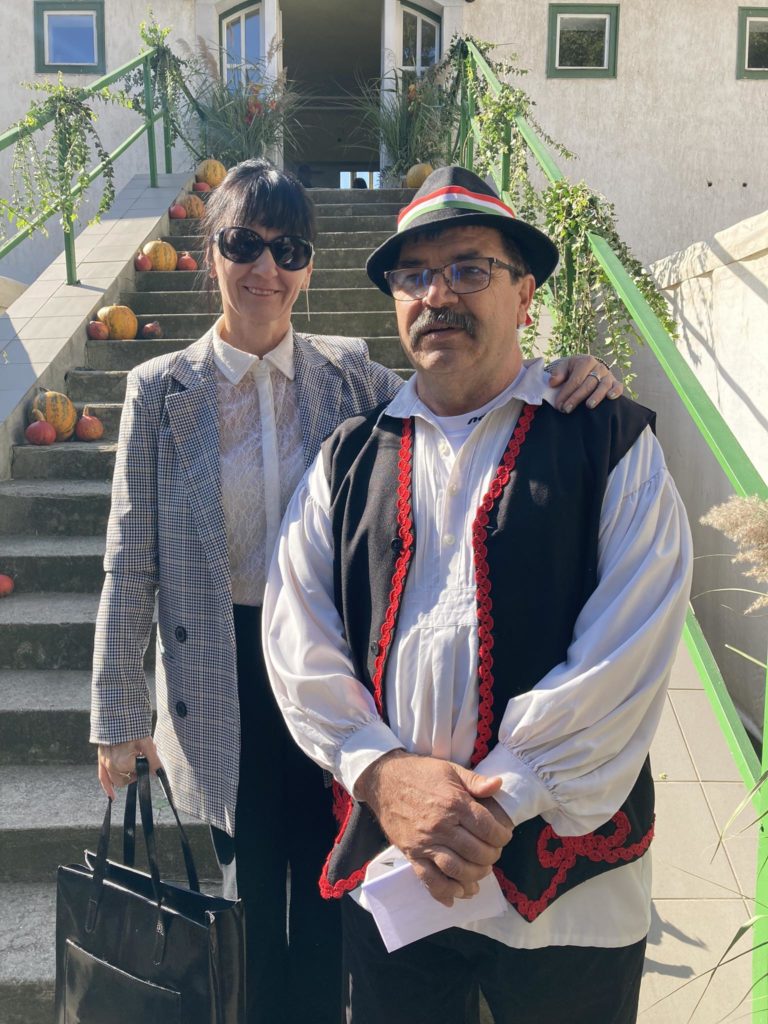 Egy napszemüveget és kockás kabátot viselő, fehér blúzt viselő nő egy hagyományos ruhába öltözött férfi mellett áll, aki piros díszítésű fehér inget, fekete mellényt és sapkát visel. A sütőtökkel és növényzettel díszített kültéri lépcső előtt állnak.