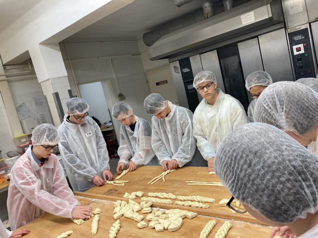 Hajhálót és fehér laborköpenyt viselő emberek csoportja összegyűlik egy fából készült asztal körül, és különféle formákat formáznak a tésztából. Úgy tűnik, hogy egy pékségben vagy konyhában vannak, rozsdamentes acél készülékekkel a háttérben.