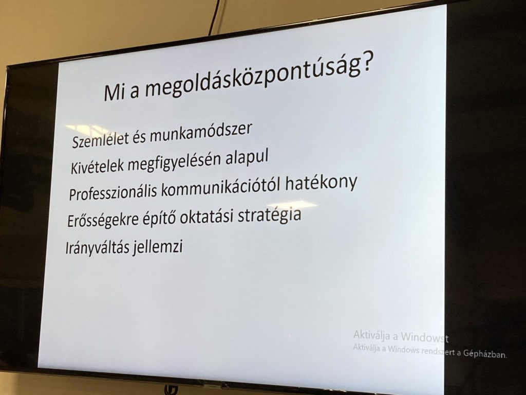 Egy prezentációs dia "Mi a megoldásközpontúság?" képernyőn jelenik meg. A dia magyar nyelven sorolja fel a pontokat, beleértve a gondolkodásmódot, a módszert, a kivételeken alapuló hatékonyságot, a hatékony szakmai kommunikációt, az erőn alapuló oktatási stratégiát és az iránymutató szempontokat.