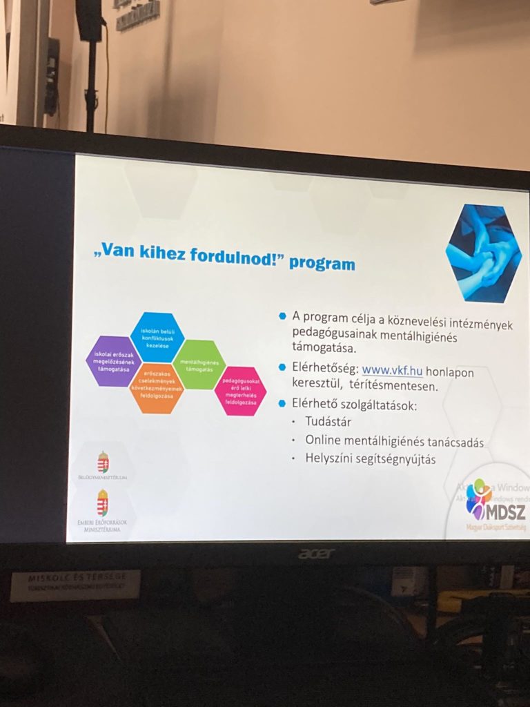 A "Van kihez fordulnod? program" című prezentációs dia a számítógép monitorán. A dián magyar nyelvű szöveg található a program céljairól és előnyeiről, valamint színes hatszögdiagram. A szervezet logói alul láthatók.