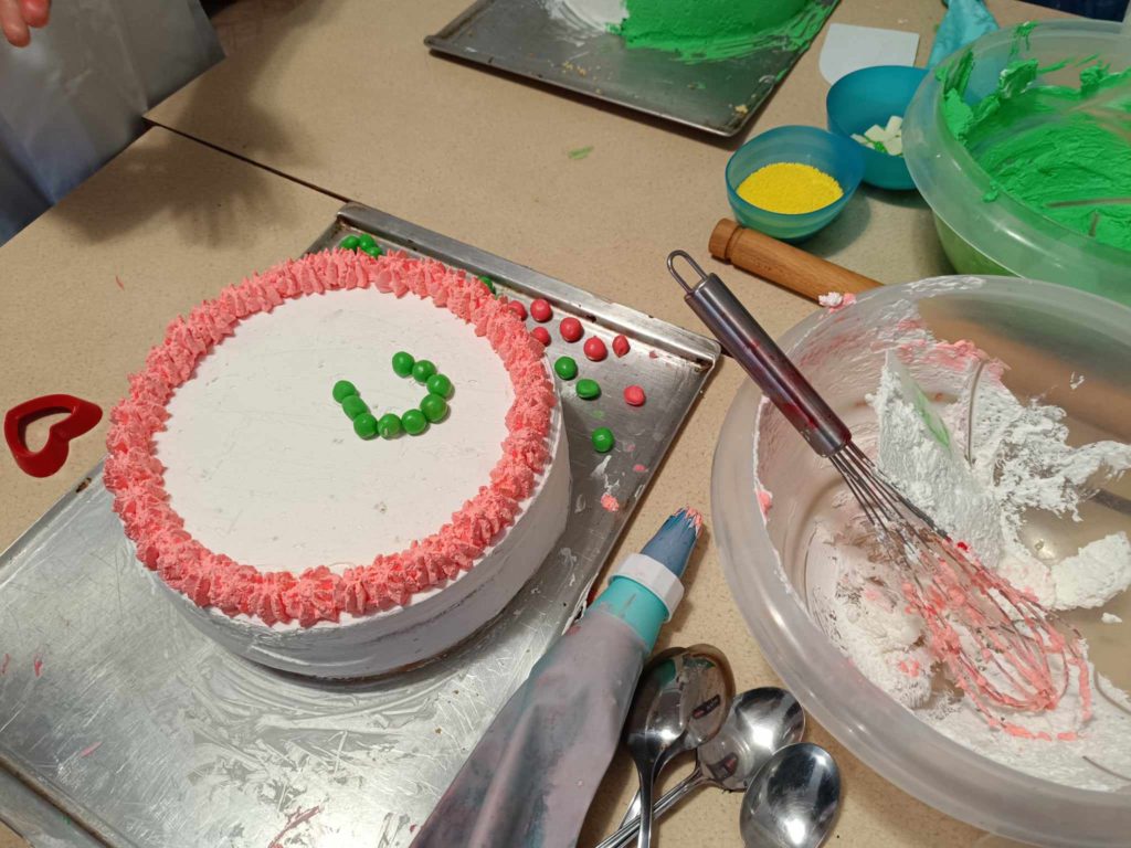 Kerek fehér torta, szélein rózsaszín cukormáz. A tortát mosolygós arcmintás zöld cukorkák díszítik. A közelben van egy fém habverő, csőszerszámok, kanalak és egy tál fehér cukormázzal, valamint zöld és sárga szórással.