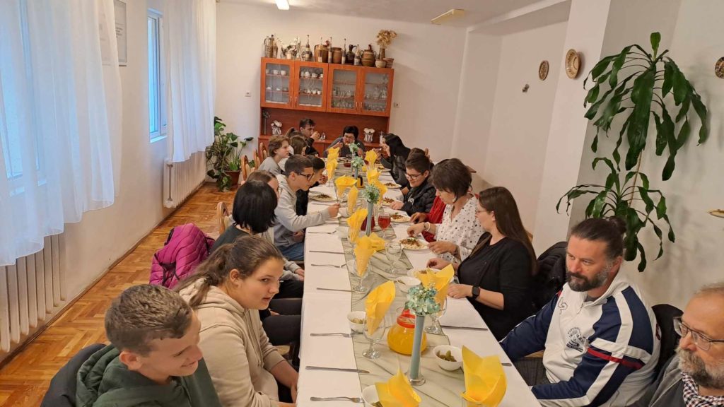 Emberek nagy csoportja, köztük felnőttek és gyerekek ülnek egy hosszú, terített étkezőasztal körül egy erősen megvilágított szobában. Az asztalt sárga szalvéták díszítik, az edények és poharak szépen el vannak rendezve. A háttérben növények és egy faszekrény látható.