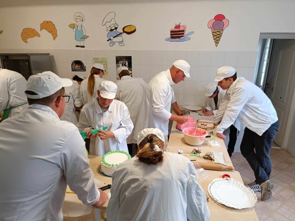Fehér kabátba és kalapba öltözött emberek csoportja dolgozik együtt a konyhában. Vannak, akik tortákat díszítenek zöld és rózsaszín cukormázzal, míg mások a munkalapokon végzett munkájukra koncentrálnak. A falakat sütemények és desszertek rajzai díszítik.