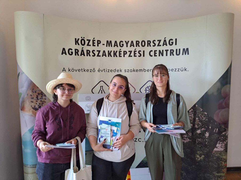 Három személy áll a Közép-Magyarországi Agrárszakképzési Centrum transzparense előtt. Brosúrákat tartanak, és mosolyognak a kamerába. A háttérben mezőgazdasági témájú grafika és szöveg található.