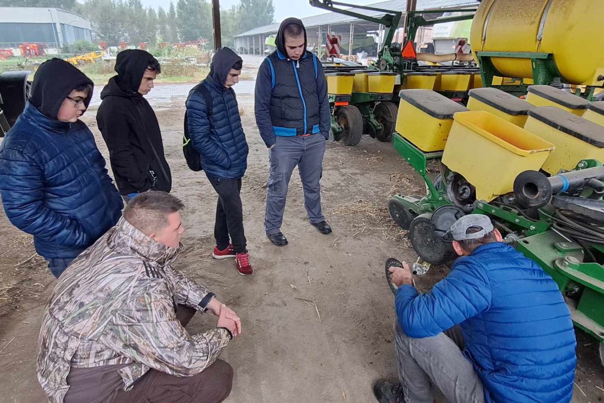 Emberek csoportja gyűlik össze egy ember körül, aki letérdel, és egy mezőgazdasági berendezésen dolgozik egy műhelyben. Egyesek kabátot és kapucnis pulcsit viselnek. A háttérben különféle gépek és egy traktor látható.