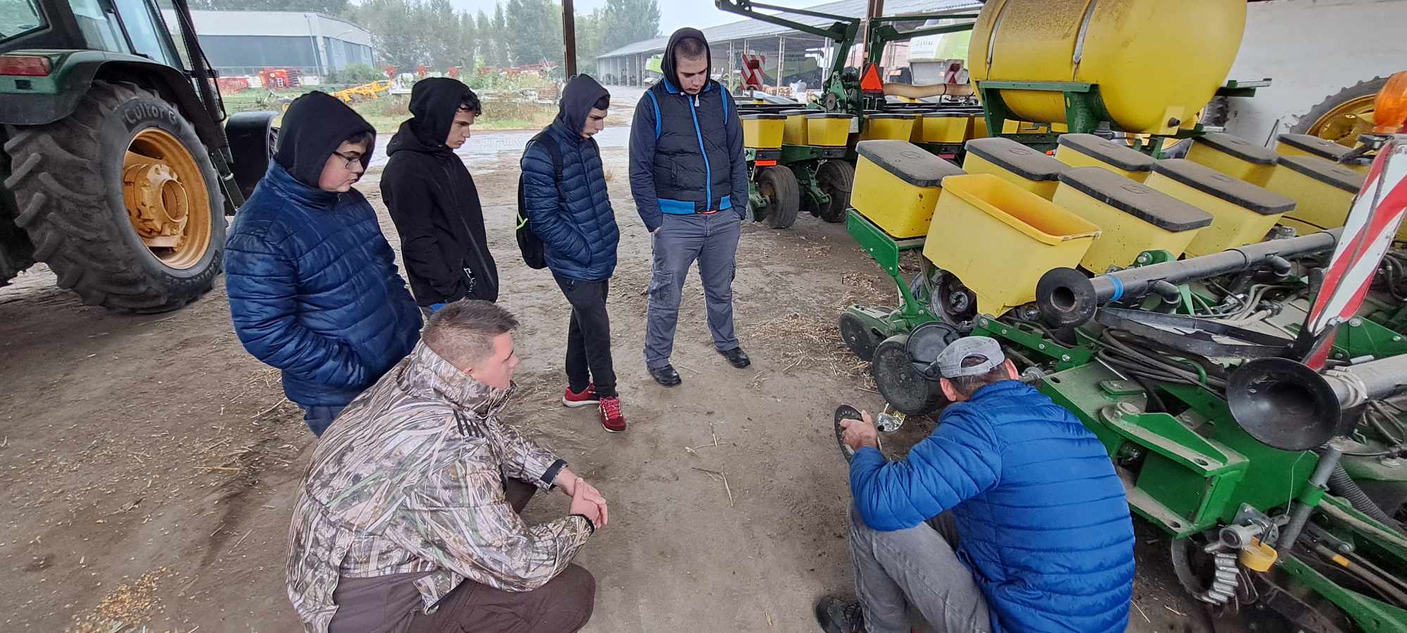Emberek csoportja gyűlik össze egy ember körül, aki letérdel, és egy mezőgazdasági berendezésen dolgozik egy műhelyben. Egyesek kabátot és kapucnis pulcsit viselnek. A háttérben különféle gépek és egy traktor látható.
