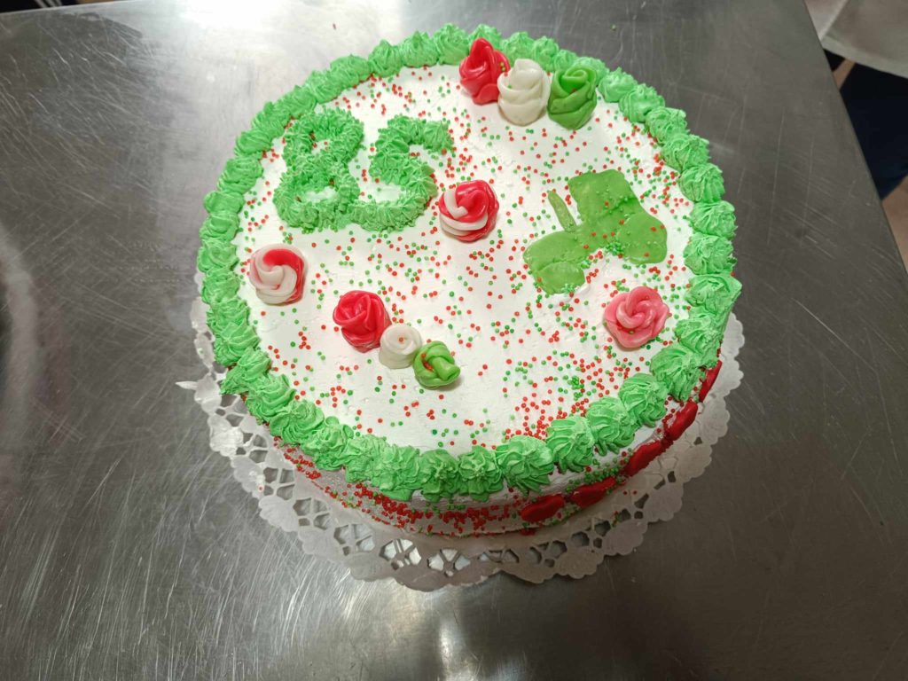 Zöld-fehér cukormázzal díszített kerek torta, tetején piros és zöld szórással, zöld és piros cukormáz rózsákkal. A tetejére zöld cukormázzal a „25” szám van írva. A torta csipkepapír szalvétán, fém felületen áll.