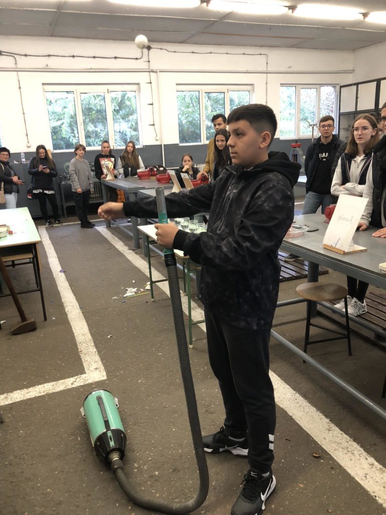 Egy fiatal fiú porszívótömlő használatát mutatja be egy osztályteremben vagy műhelyben, miközben egy csoport diák és felnőtt figyeli. Különféle eszközök, anyagok és projektek láthatók az asztalokon a szobában.