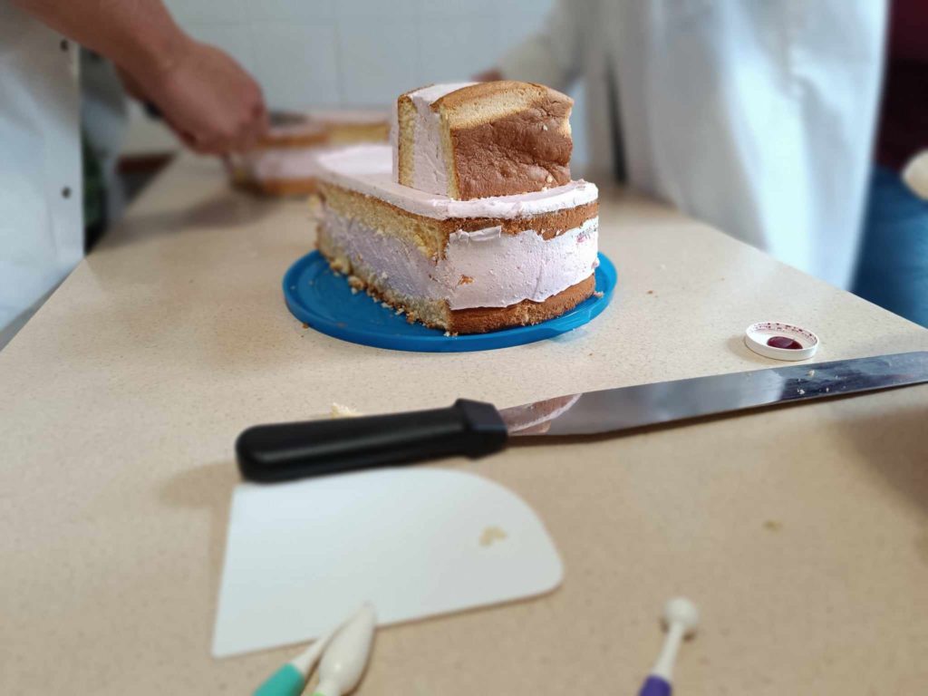 Egy részlegesen faragott torta rózsaszín cukormázzal egy kék tálcán ül a pulton. Egy nagy kés, egy kis műanyag kaparó és néhány díszítőeszköz van elszórva a közelben. Két ember dolgozik a háttérben, készítik vagy díszítik a tortát.