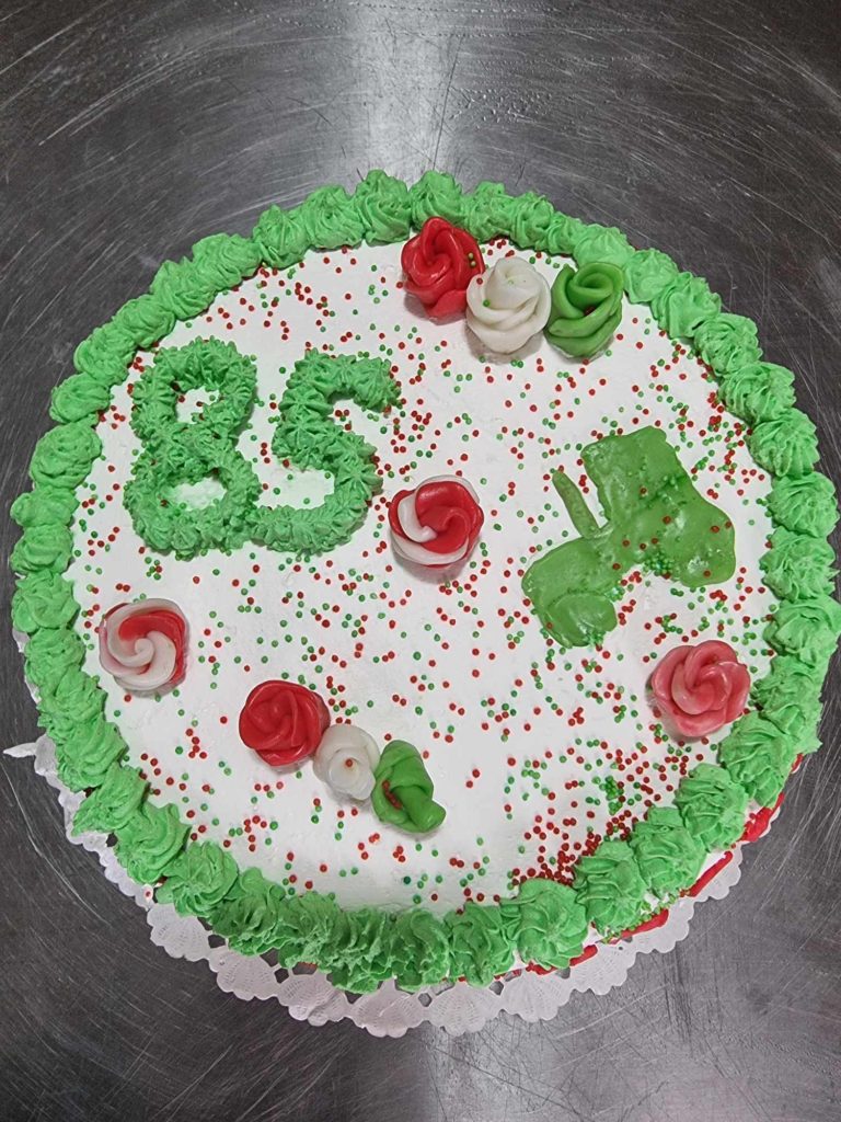 Zöld, fehér és piros cukormázzal díszített kerek torta. A tortán zöld cukormázzal a „85” szám van írva, a tetejére pedig piros, zöld és fehér rózsa alakú díszítések, szórványok kerültek.