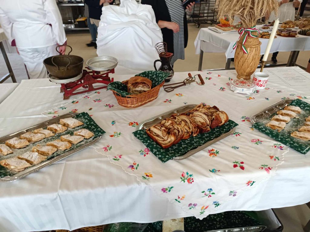 A virágokkal díszített fehér terítős asztalon különféle sütemények, köztük rétesek és fonott kenyér látható. Van még búza díszítés vázában és hagyományos kávédaráló. A háttérben fehér ruhás emberek láthatók.