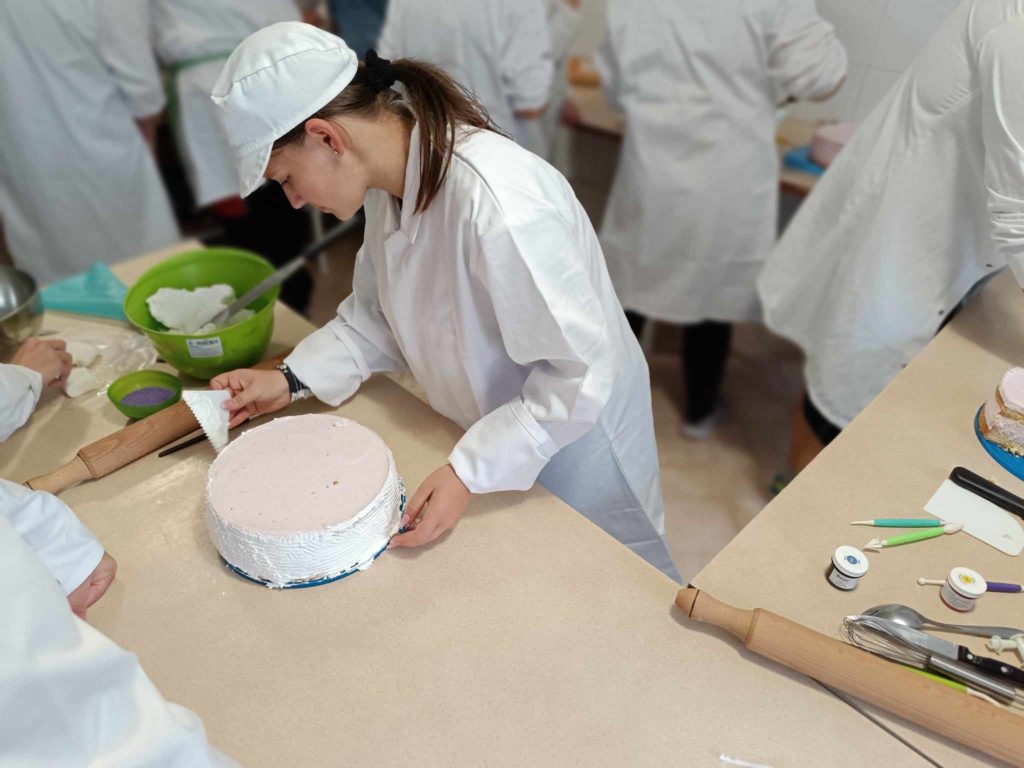 Egy fehér kabátot és sapkát viselő személy egy spatula segítségével egy kerek tortát díszít fehér cukormázzal. Mások is körülveszik őket hasonló öltözékben konyhai környezetben, a pulton láthatóak a különféle sütőeszközök és hozzávalók.