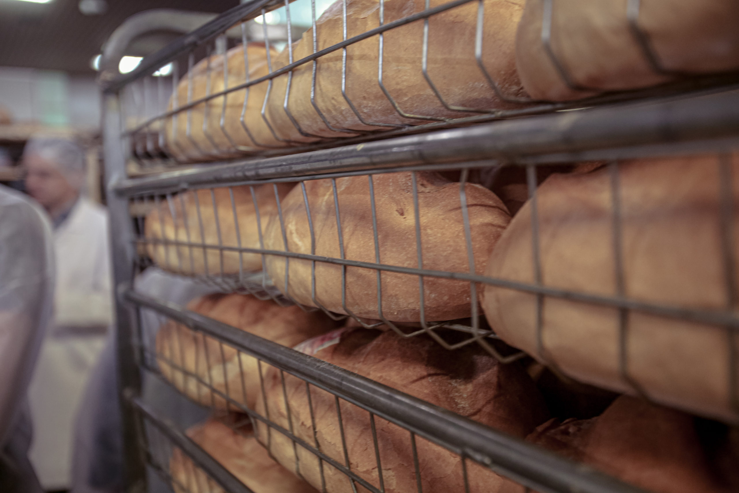Frissen sült kenyérrel töltött állvány egy pékségben. A kenyerek több polcon szépen egymásra vannak rakva. A háttérben fehér egyenruhás munkások láthatók, kissé életlenül.