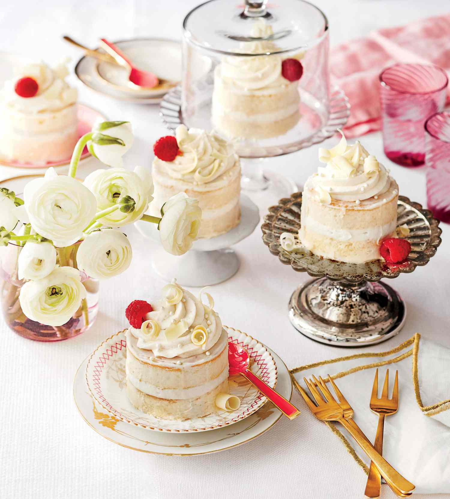 Egy gyönyörűen megterített asztalon egyedi kis sütemények láthatók tejszínhabbal és málnával. Egyes sütemények díszes állványokon, míg mások üveglapokban vannak. Az asztalon fehér ranunculus virágok is találhatók vázában, színes poharak és arany edények.