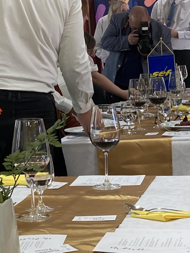 Emberek csoportja összegyűlik egy hivatalos eseményre terített asztal körül. Az asztalt fehér terítők, arany futók, borospoharak és virágdíszek díszítik. A háttérben egy fotós örökíti meg a pillanatot.