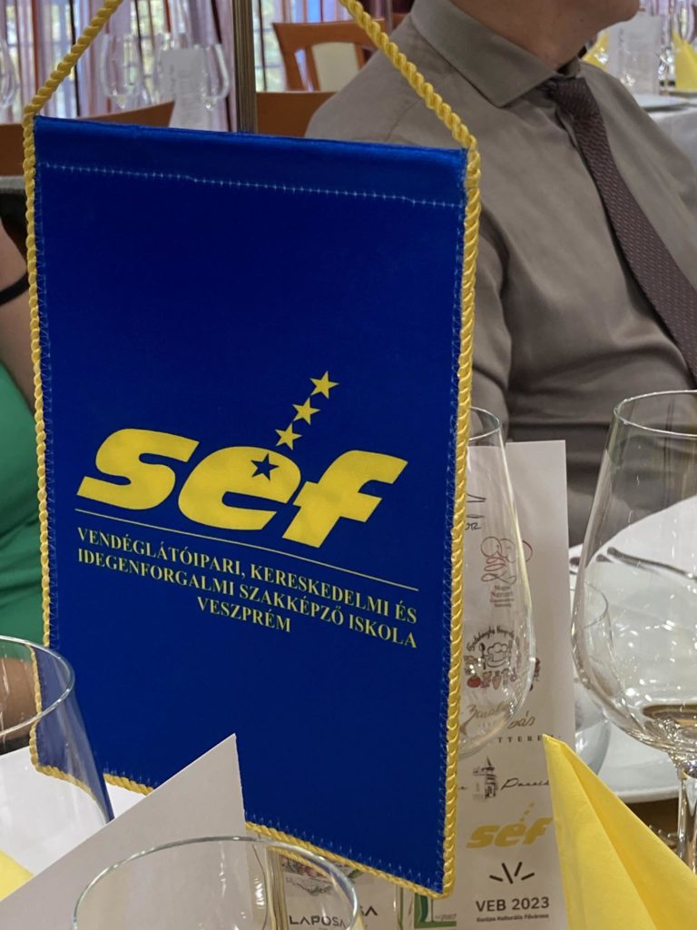 Az étkezőasztalon egy kék színű, sárga feliratú transzparens látható. A transzparensen a "SEF" felirat olvasható, alatta további magyar szöveggel. A transzparens körül több borospohár, egy összehajtott sárga szalvéta és a háttérben étkezők láthatók.