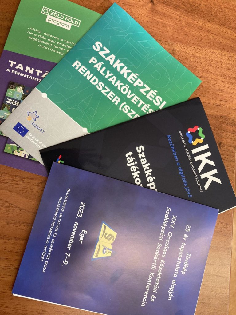 Egy asztallapon öt színes magyar nyelvű füzet látható. A füzetek különféle szövegeket és logókat jelenítenek meg, köztük az Európai Unió zászlaját. Az oktatási és szakképzési programokra összpontosítanak, láthatóak a dátumok és a konferencia információi.