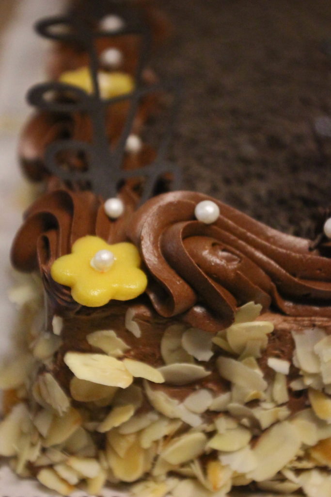 Közeli kép egy csokoládé örvényekkel, sárga virág alakú fondant díszítéssel és fehér cukorgyöngyökkel díszített csokitorta sarkáról. Az oldalát szeletelt mandulával borítjuk.