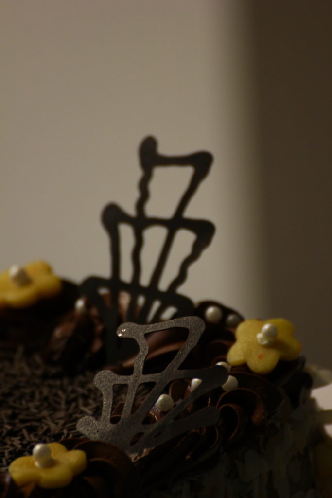 Egy csokoládé torta tetejének közeli képén étcsokoládé absztrakt formák és kis sárga virágdíszek láthatók fehér középponttal. A háttér elmosódott, a figyelem a bonyolult és művészi tortafeltétekre összpontosul.