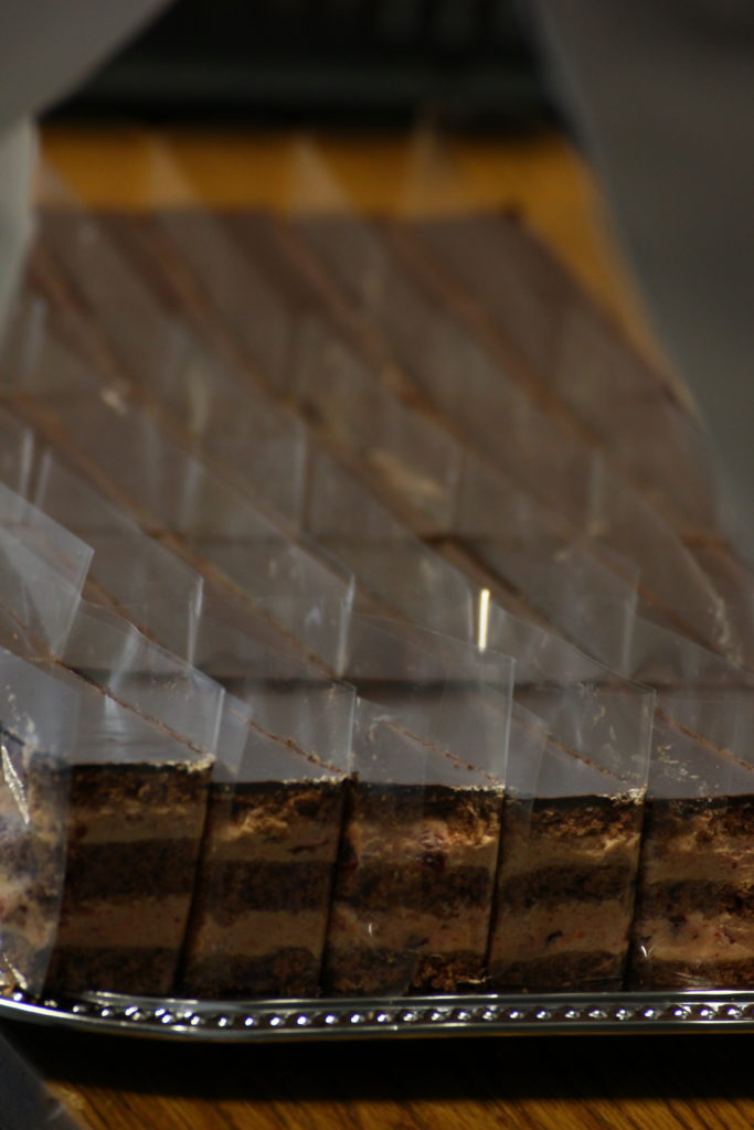 Egy tálca szépen elrendezett, egyenként csomagolt csokitortákból. A tortákat téglalap alakú darabokra szeleteljük, és az átlátszó csomagoláson keresztül láthatók a rétegek. A tálcát fa felületre helyezzük.