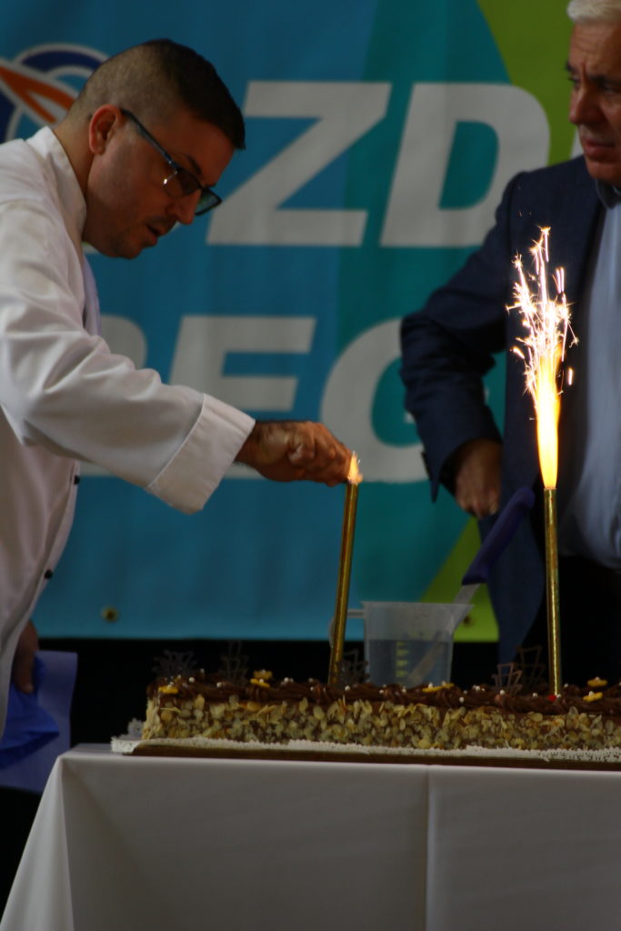 Egy fehér köpenyes ember csillagszórót gyújt egy nagy, téglalap alakú tortán, amelyet dióval és csokoládédíszek díszítenek. Egy másik kék kabátos ember áll a közelben. A háttérben színes banner látható részben látható szöveggel.