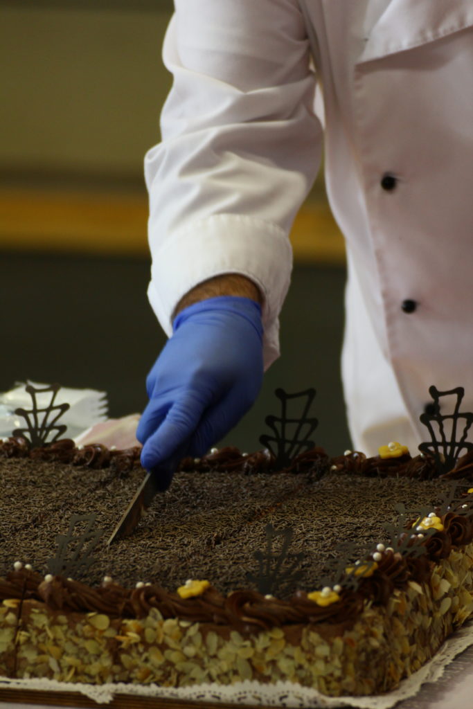 Egy fehér köpenyt és kék kesztyűt viselő szakács nagy, téglalap alakú, fogazott szélű csokitortát vág. A tortát csokoládéreszelékkel, oldalán mandulaszeletekkel, tetején bonyolult csokoládé díszítéssel díszítjük.