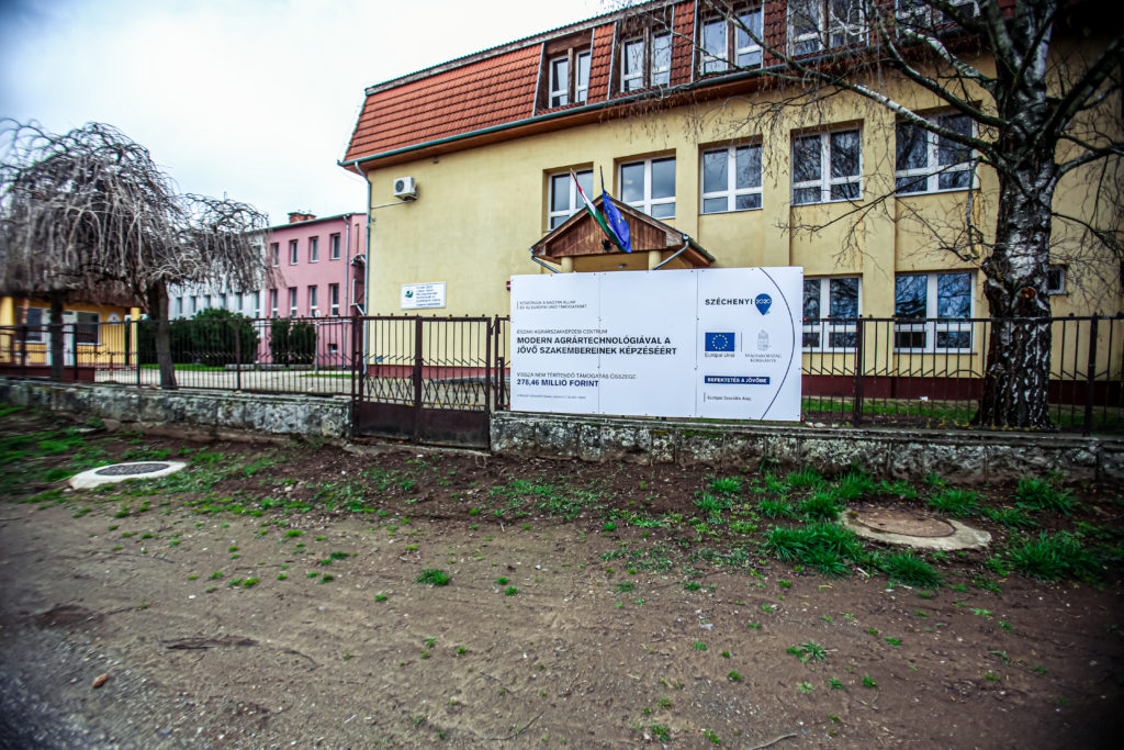 Egy vörös cseréptetős, sárga külsővel fémkerítés mögött áll az épület. Az épület előtt egy nagy fehér tábla látható a fejlesztési projektekkel kapcsolatos információkkal, beleértve az Európai Unió és a helyi hatóságok logóit. A jobb oldalon egy kis fa áll.