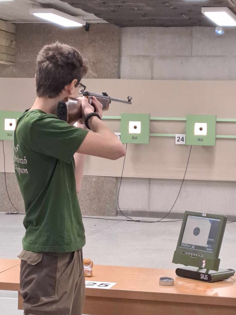 Egy rövid hajú, szemüveges és zöld inget viselő személy puskával céloz egy fedett lőtéren. A célpont egy „24” feliratú zöld táblára van felszerelve, a célpont nézetét megjelenítő monitor pedig egy közeli faasztalon van.
