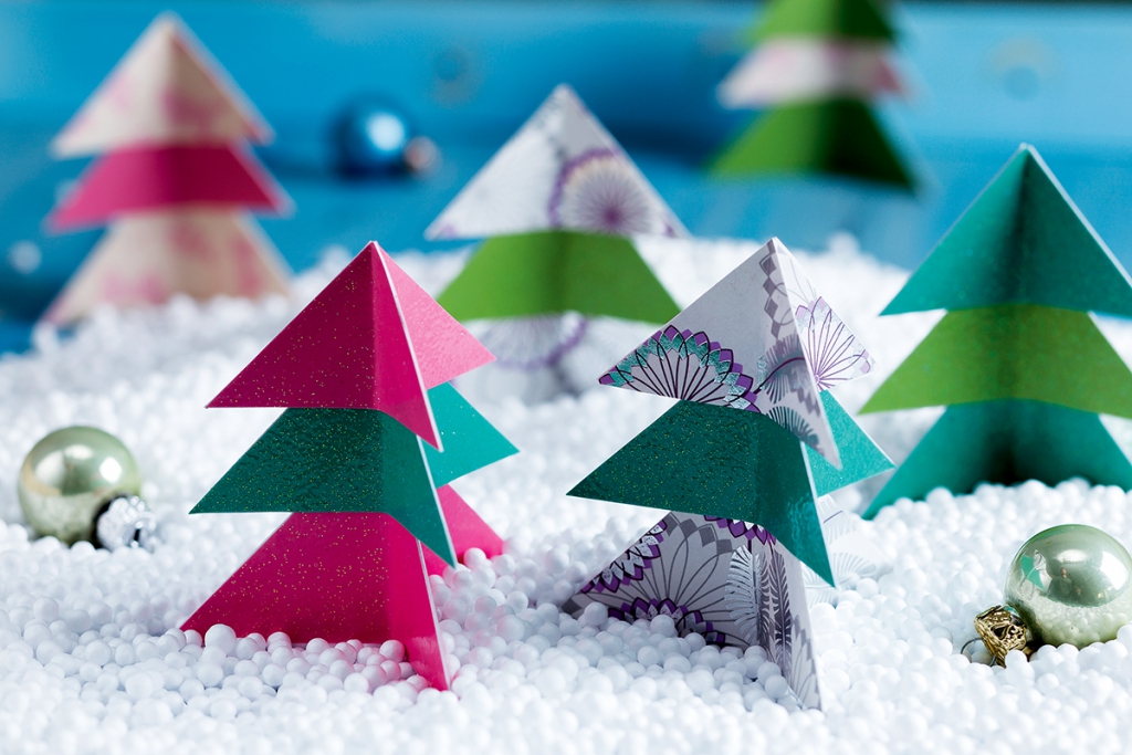 Ünnepi jelenet színes, rózsaszín, zöld és mintás papírból készült origami karácsonyfákkal, amelyek a hóhoz hasonlító fehér hungarocell gyöngyökből álló ágyon állnak. A háttérben kék felületen csillogó díszek sorakoznak.