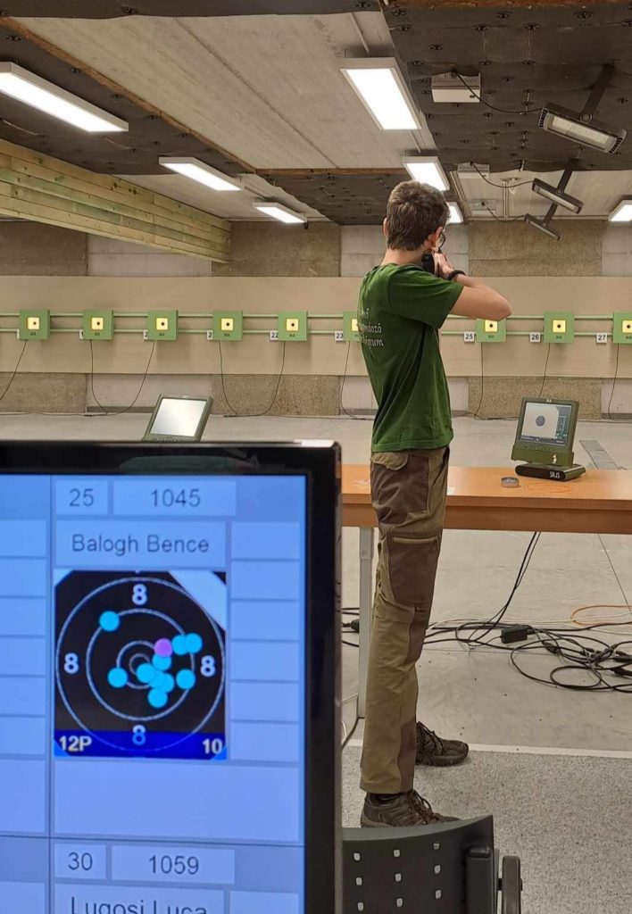 Zöld ingbe és barna nadrágba öltözött személy pisztollyal céloz egy fedett lőtéren. Az előtérben egy monitor látható, amin egy célpont látható "Balogh Bence" és "Lugosi Luca" felirattal.