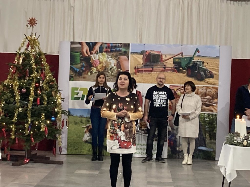 Egy nő áll és beszél a bal oldalon karácsonyfával feldíszített színpadon, jobb oldalon pedig gyertyákat gyújtanak. Mögötte három ember áll egy mezőgazdasági képeket megjelenítő háttér előtt.