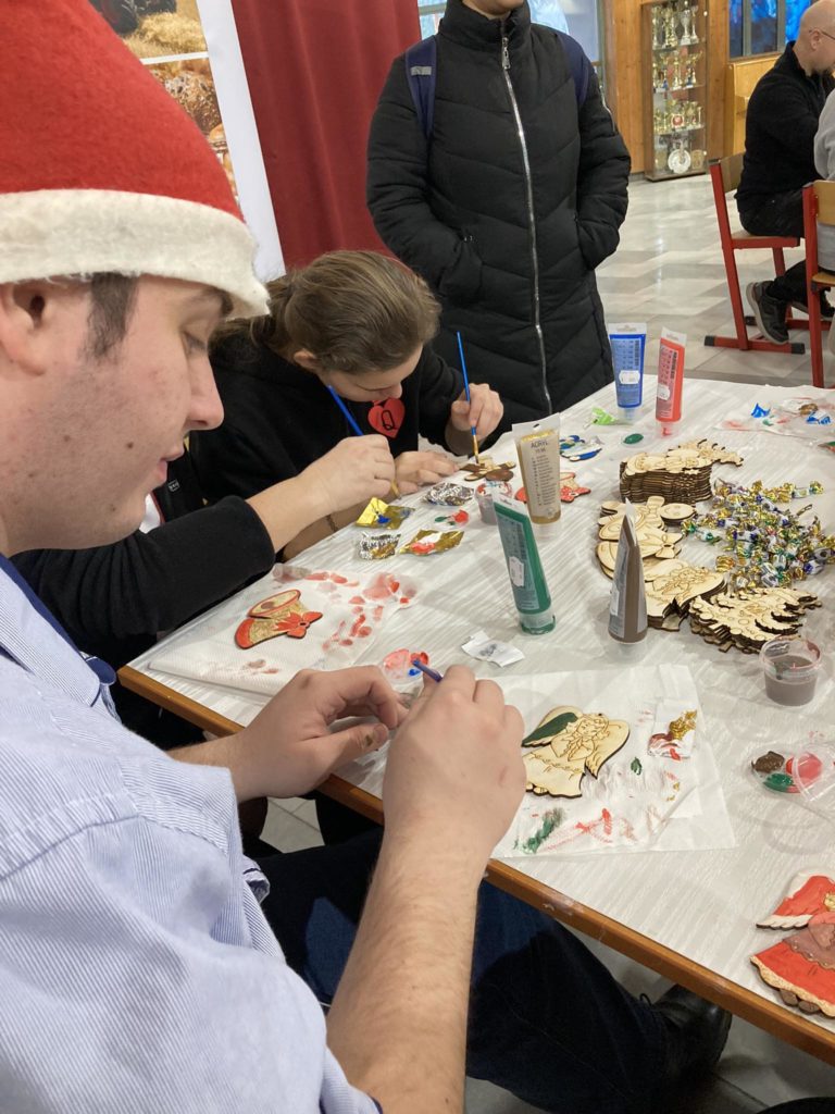Egy Mikulás sapkás férfi és két kabátos ember ül egy asztalnál, és fából készült karácsonyi díszeket fest. Különböző festékek és festett dísztárgyak vannak szétszórva az asztalon. A háttérben ünnepi díszek és egy álló személy látható.