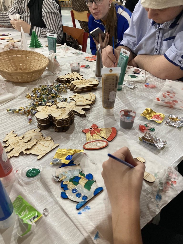 Az emberek egy asztal körül gyűlnek össze, amelyet különféle kézműves kellékekkel, köztük festékkel, fadíszekkel és csillámokkal díszítettek. Úgy tűnik, ünnepi témájú díszeket festenek és díszítenek.