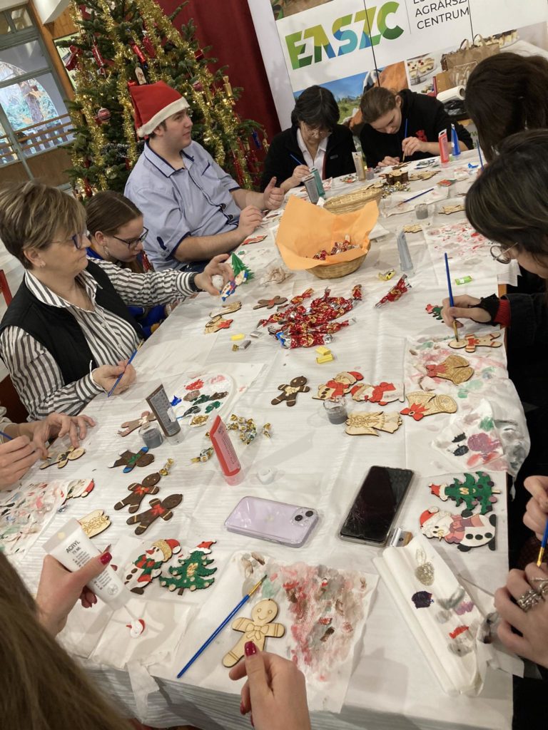 Emberek csoportja gyűlt össze egy nagy asztal körül, és aktívan foglalkozott karácsonyi kézművességgel. Az asztalt ünnepi díszek, ragasztó és festék borítják. A háttérben egy feldíszített karácsonyfa és egy „EASZC” feliratú transzparens látható.