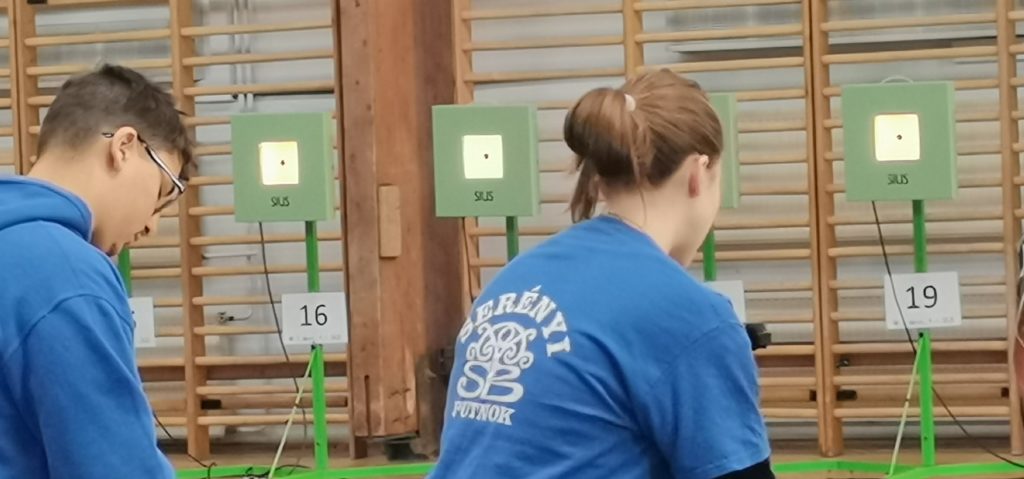 Két személy vesz részt egy lőtéri tevékenységben, mindketten kék inget viselnek. A jobb oldali személy hátán egy logó található "SB" szöveggel. A 16-os és 19-es célszámok láthatók a háttérben.