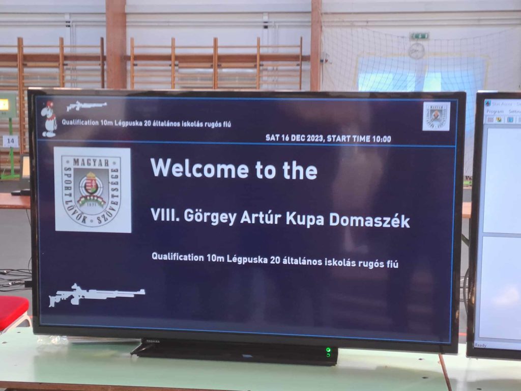 Egy tévéképernyő, amely információkat jelenít meg a VIII. Görgey Artúr Kupa Domaszéken. A rendezvény egy 10 méteres légpuska verseny általános iskolás fiúknak 2023. december 16-án 10:00 órakor.